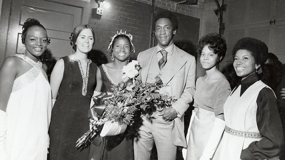 Five female members of the 1969 Homecoming Court are pictured with Bill Cosby. The Homecoming Queen, Carolyn Williams, is wearing a large tiara and holding a bouquet of roses. 