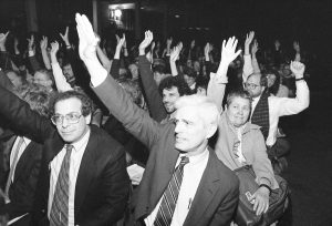 Several dozen people seated in a room, hands raised in support of the vote to remove the ROTC from campus. In the foreground is Professor Joe Elder.