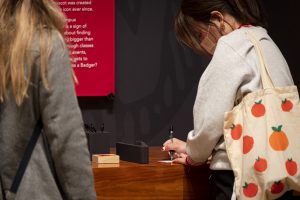 A woman with a gray sweatshirt and tote bag stands and writes with a pen on a wooden counter.