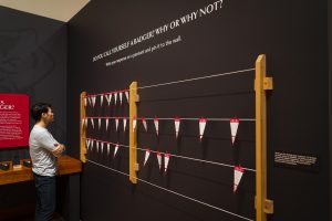 A man win a gray shirt stands in front of a black wall where red and white pennants hang on horizontal strings.