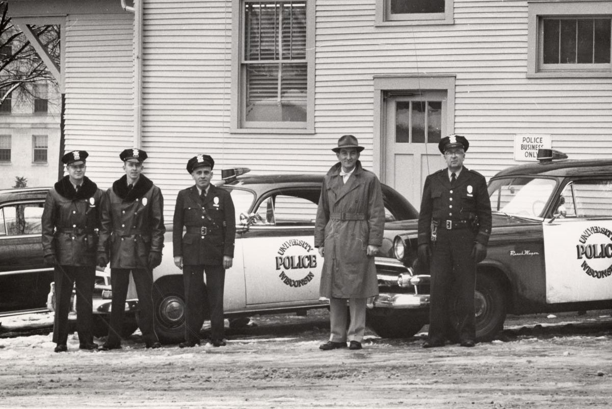 Four men in police uniforms and one man in a trench coat stand on a snowy street in front of two police cars.