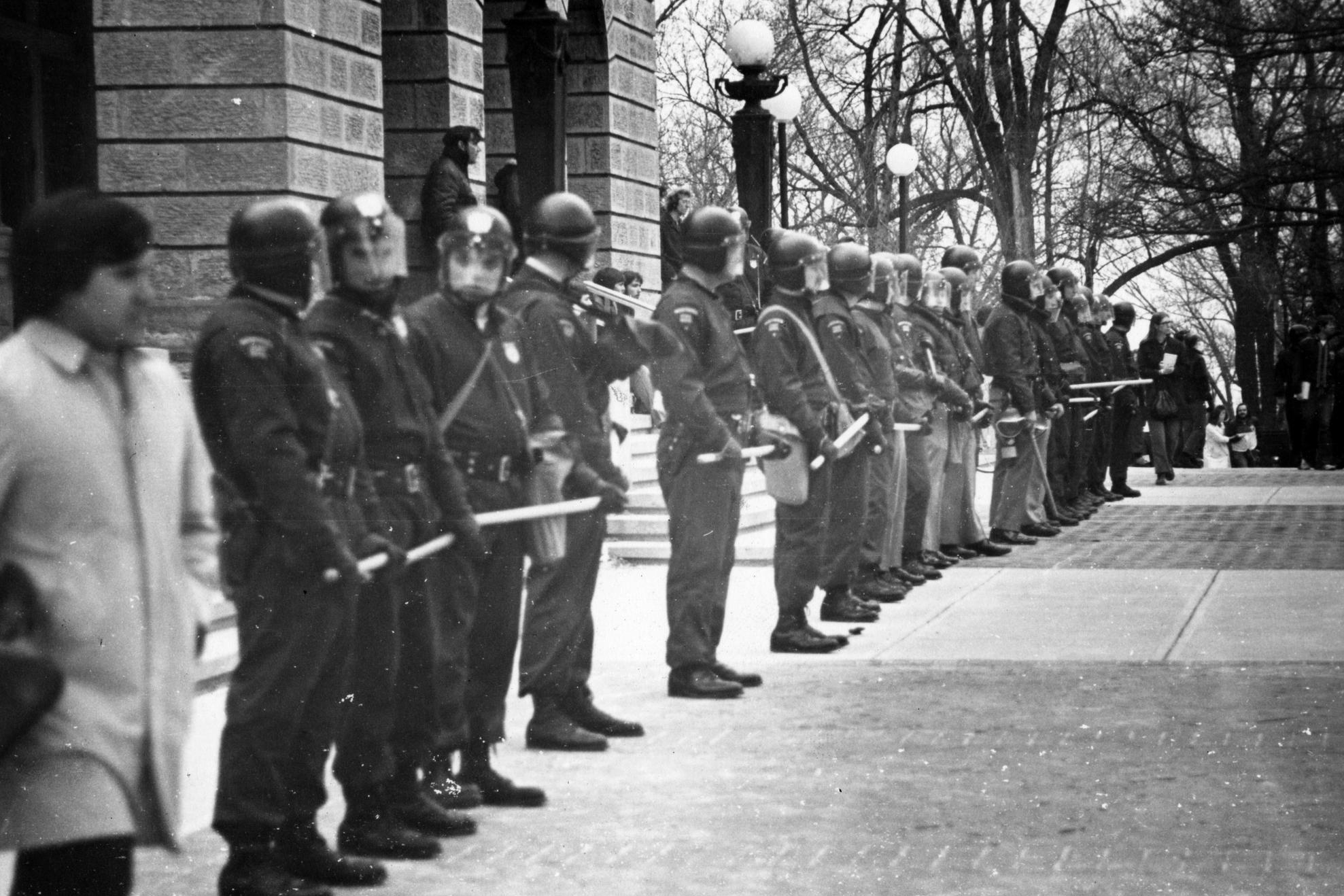Several police officers wearing dark uniforms and riot helmets stand shoulder to shoulder in a line in front of Bascom Hall