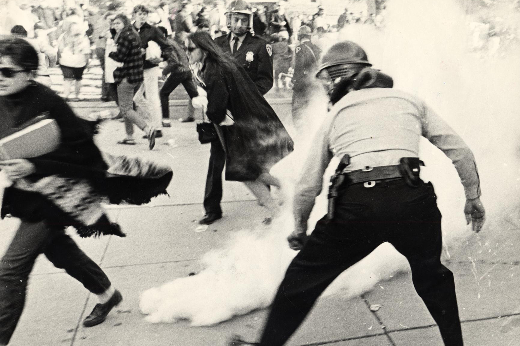 Several people run away from a cloud of tear gas. A police officer wears a gas mask on his face in the foreground.