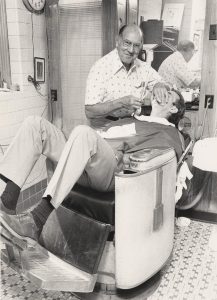 Black and white photo shows a bald man in a white shirt shaving the face of a person leaning back in a barber's chair.