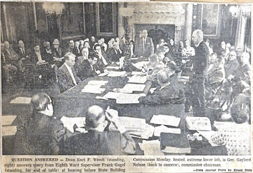 Black and white newspaper photo shows dozens of people in a large meeting room with a large wooden table in the center.