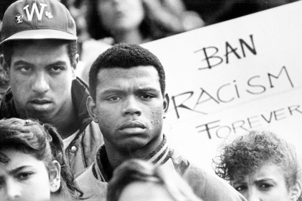 Close-up of several demonstrators a at anti-racism protest with a sign in background that reads, "Ban Racism Forever."