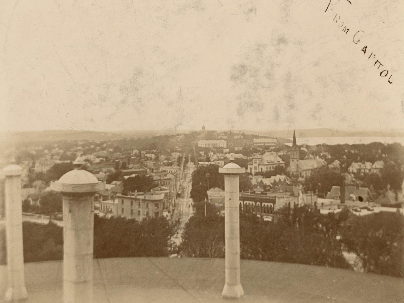 A sepia-toned, high-angle historical photograph looking down a long city street toward a distant dome building, captured from a rooftop or tower with architectural pillars visible in the foreground.