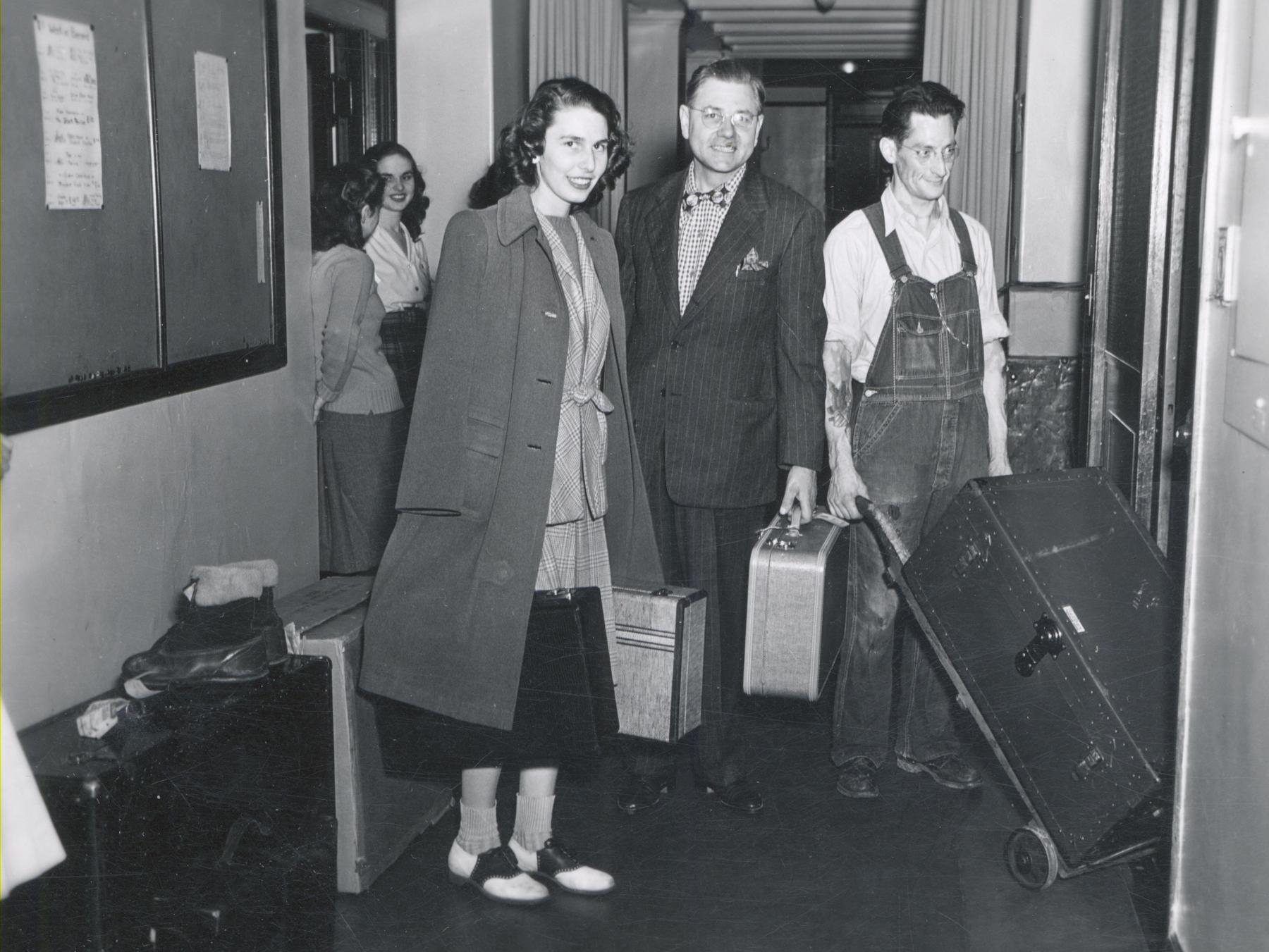 A black-and-white historical photograph of a smiling young woman in a winter coat and a man in a pinstripe suit carrying suitcases down a hallway, accompanied by a worker in overalls wheeling a large trunk on a hand truck.