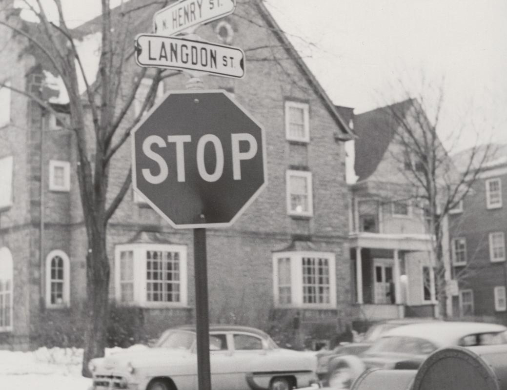 A vintage black-and-white photograph focusing on a street sign for 'Langdon St.' and a stop sign, with historic multi-story houses and mid-century cars blurred in the background.