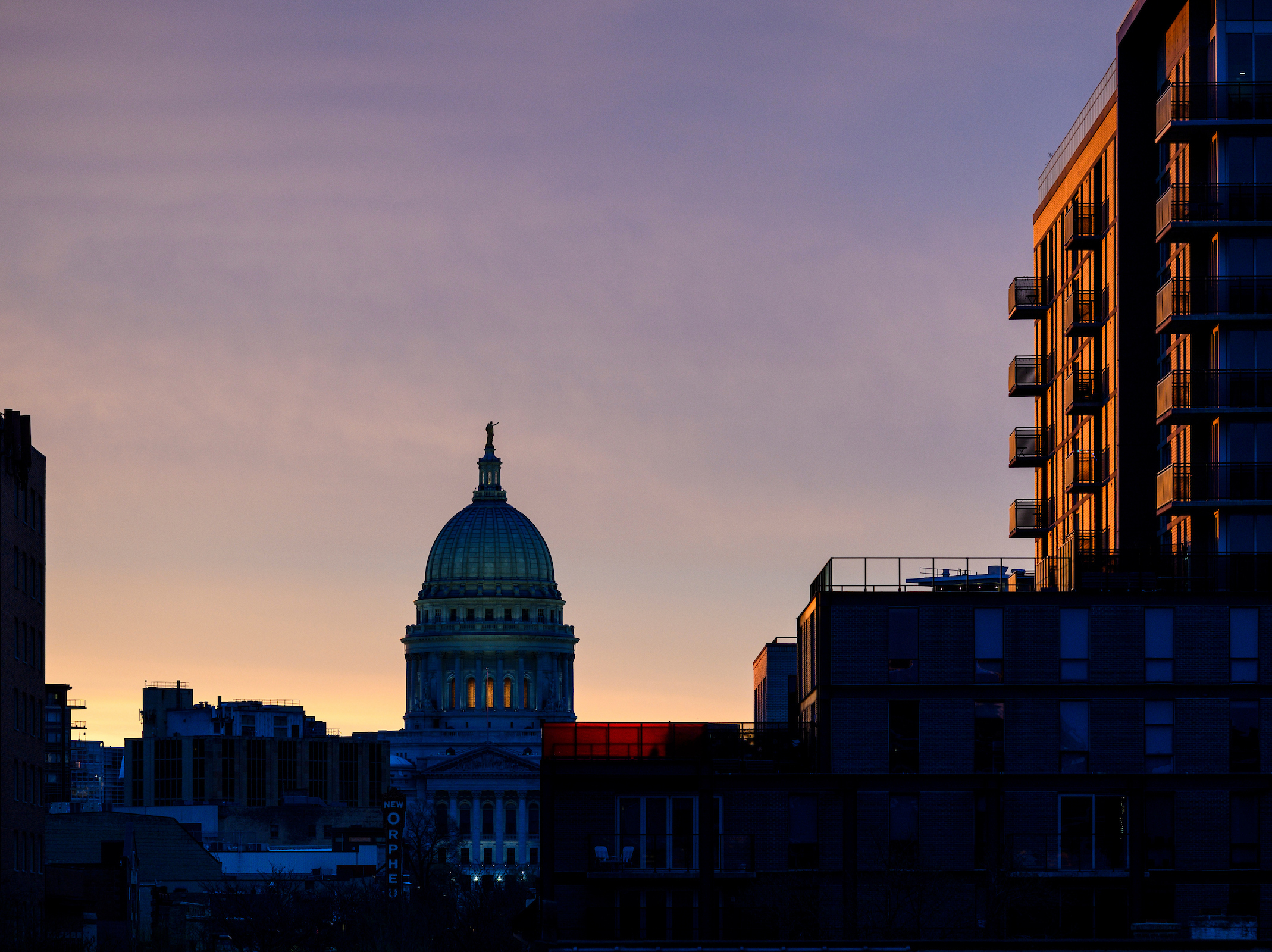 A skyline view at dusk showing the illuminated dome of the Wisconsin State Capitol building flanked by dark silhouettes of modern city buildings against a gradient purple and orange sky.
